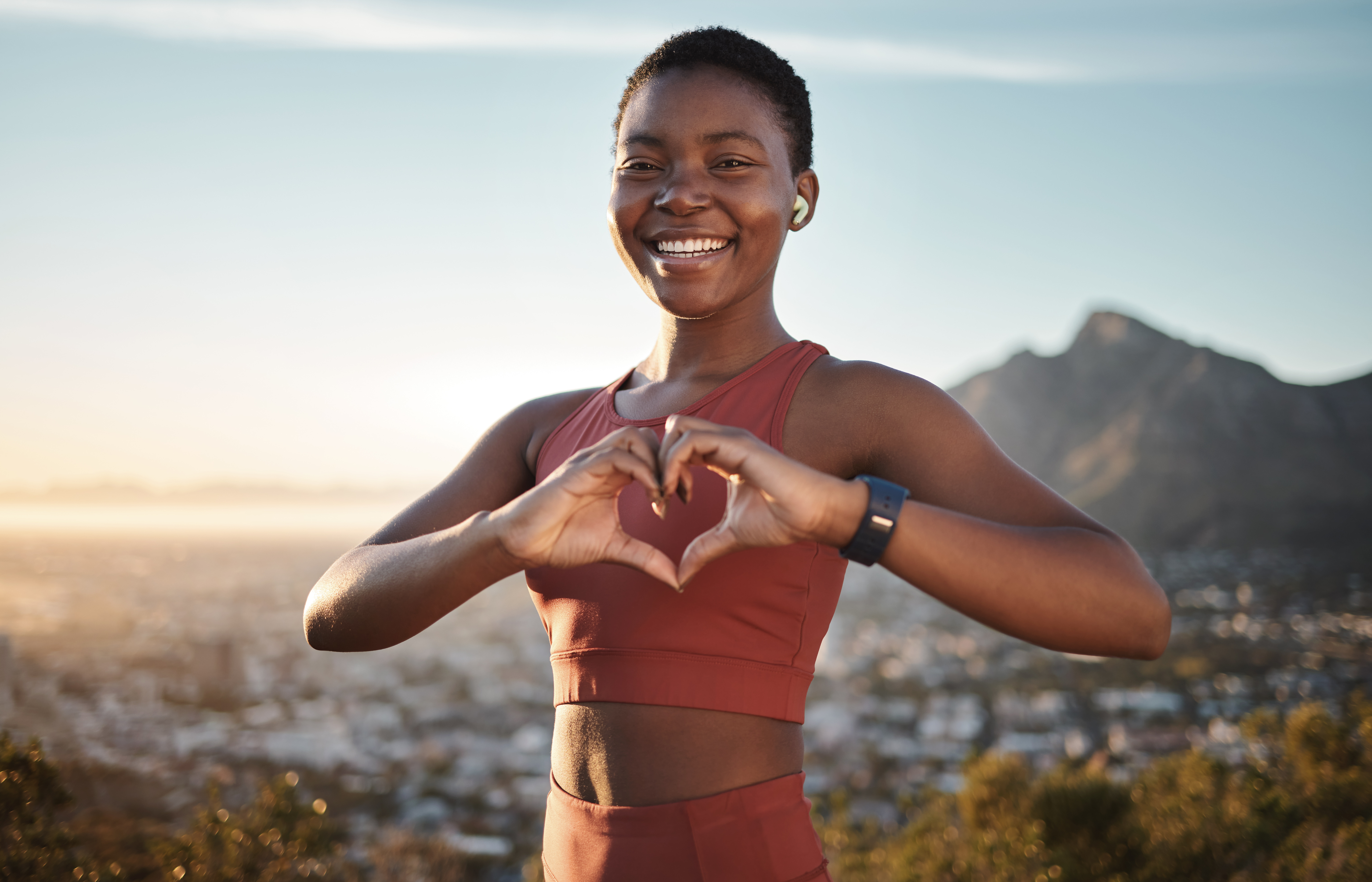 Smiling person in activewear forms a heart with hands, mountain backdrop.
