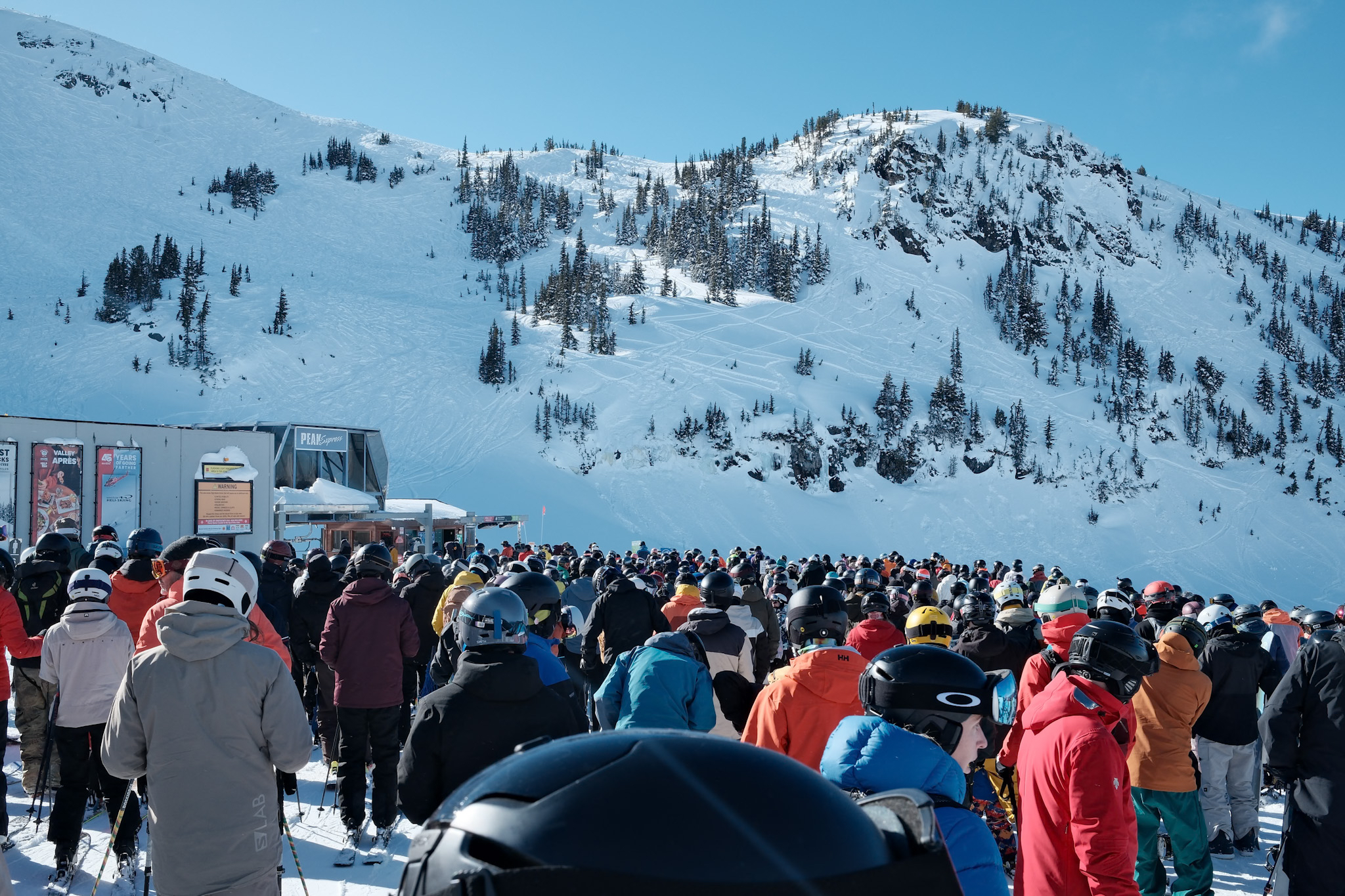 Skiers in colorful jackets gathered at snowy mountain base under clear blue sky.
