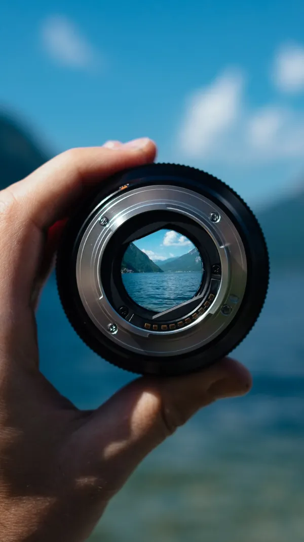 Camera lens held in hand with mountain view through it, bright blue sky.