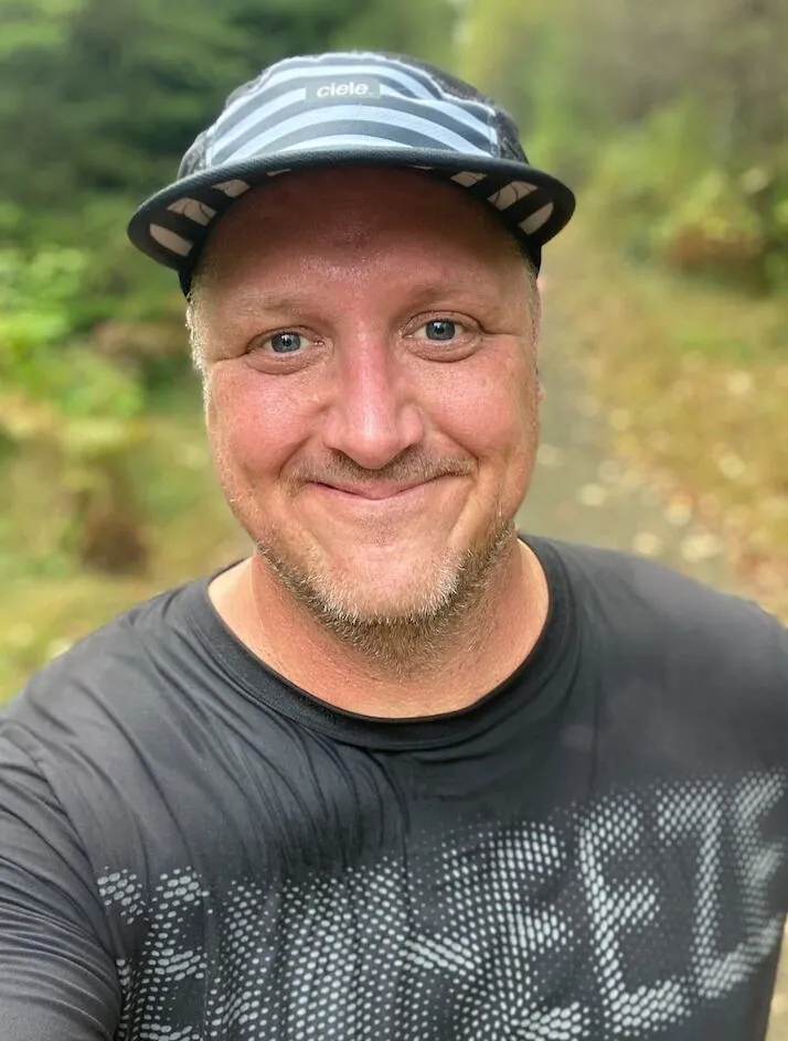 Man smiling outdoors, wearing a cap and black shirt, on a wooded path.