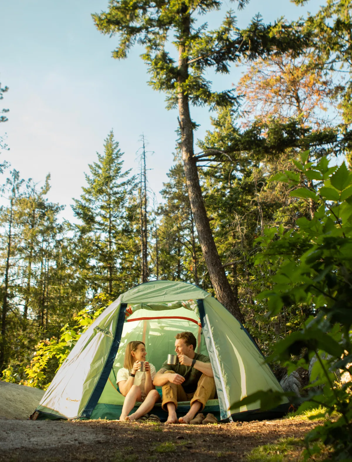 Couple sitting at a tent entrance in a sunlit forest.