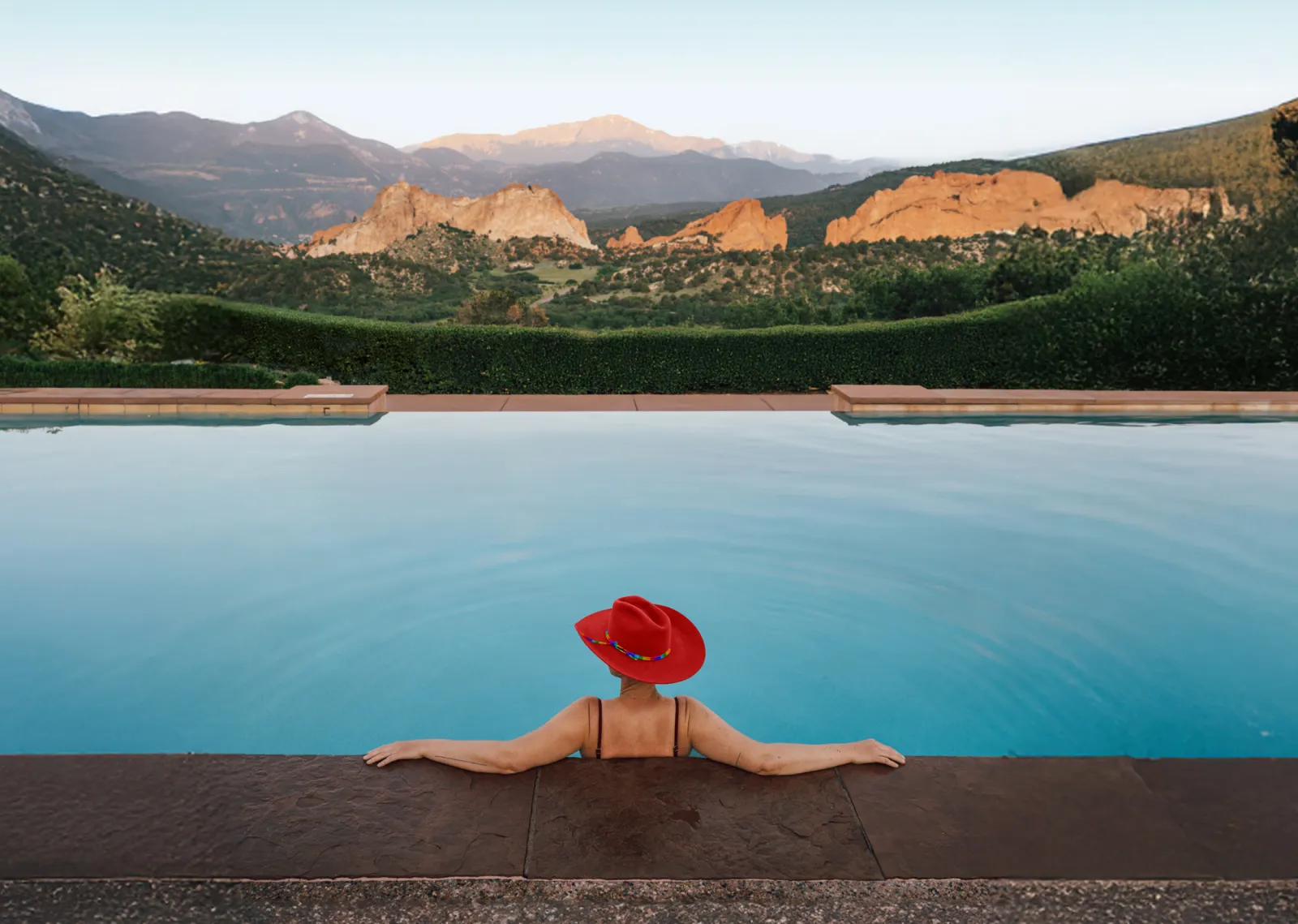 Person in a red hat relaxes in a pool, overlooking mountains at sunset.