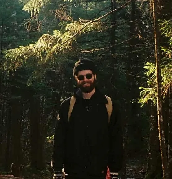 Man in a forest wearing a dark jacket and sunglasses.