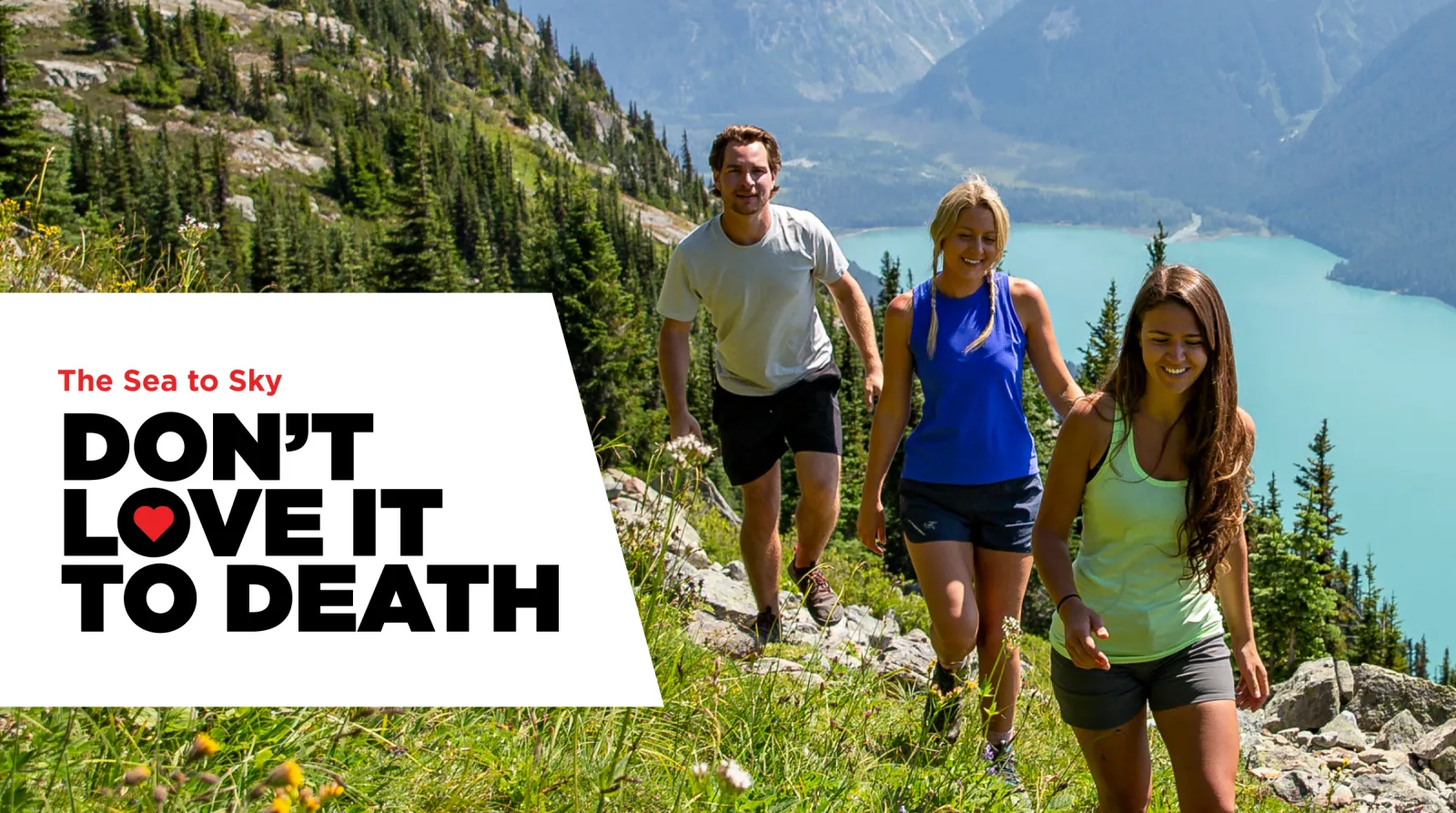 Three people hiking on a grassy mountain trail with a lake in the background.