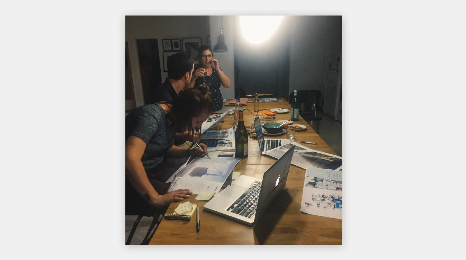 A photo of three people proofing a magazine at night with a bottle of wine