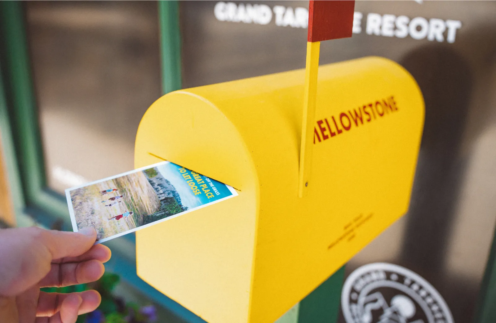 Hand inserting a postcard into yellow mailbox with "Mellowstone" text.
