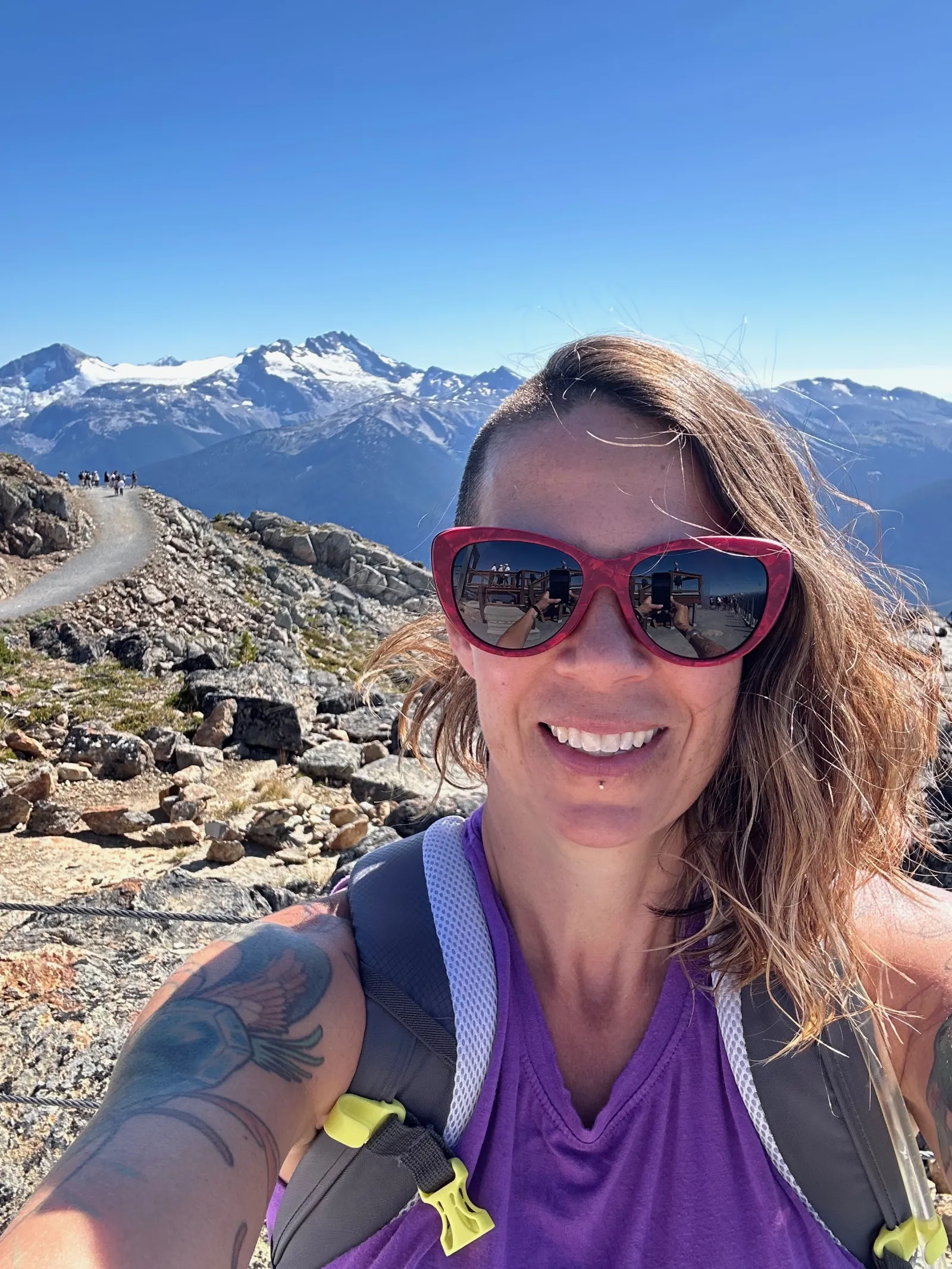 Woman wearing sunglasses smiles on a sunny mountain trail.