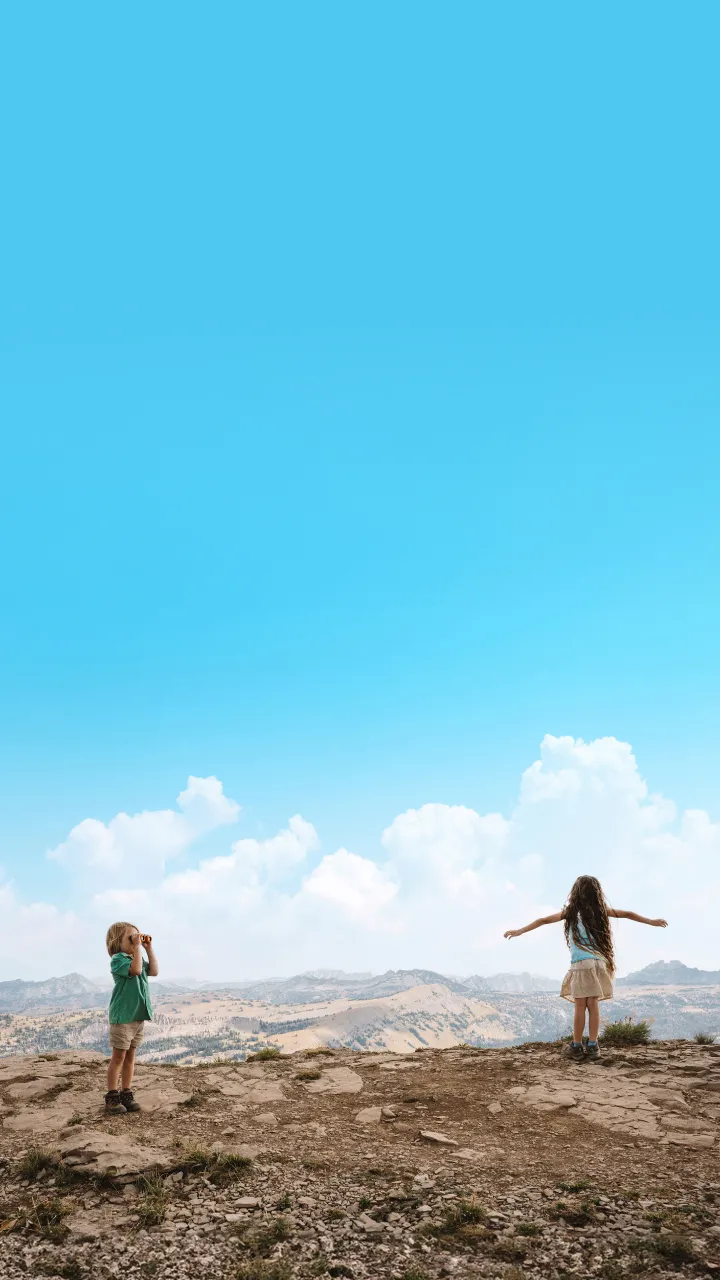 Two children on a rocky hilltop, one with arms outstretched, under a bright blue sky.