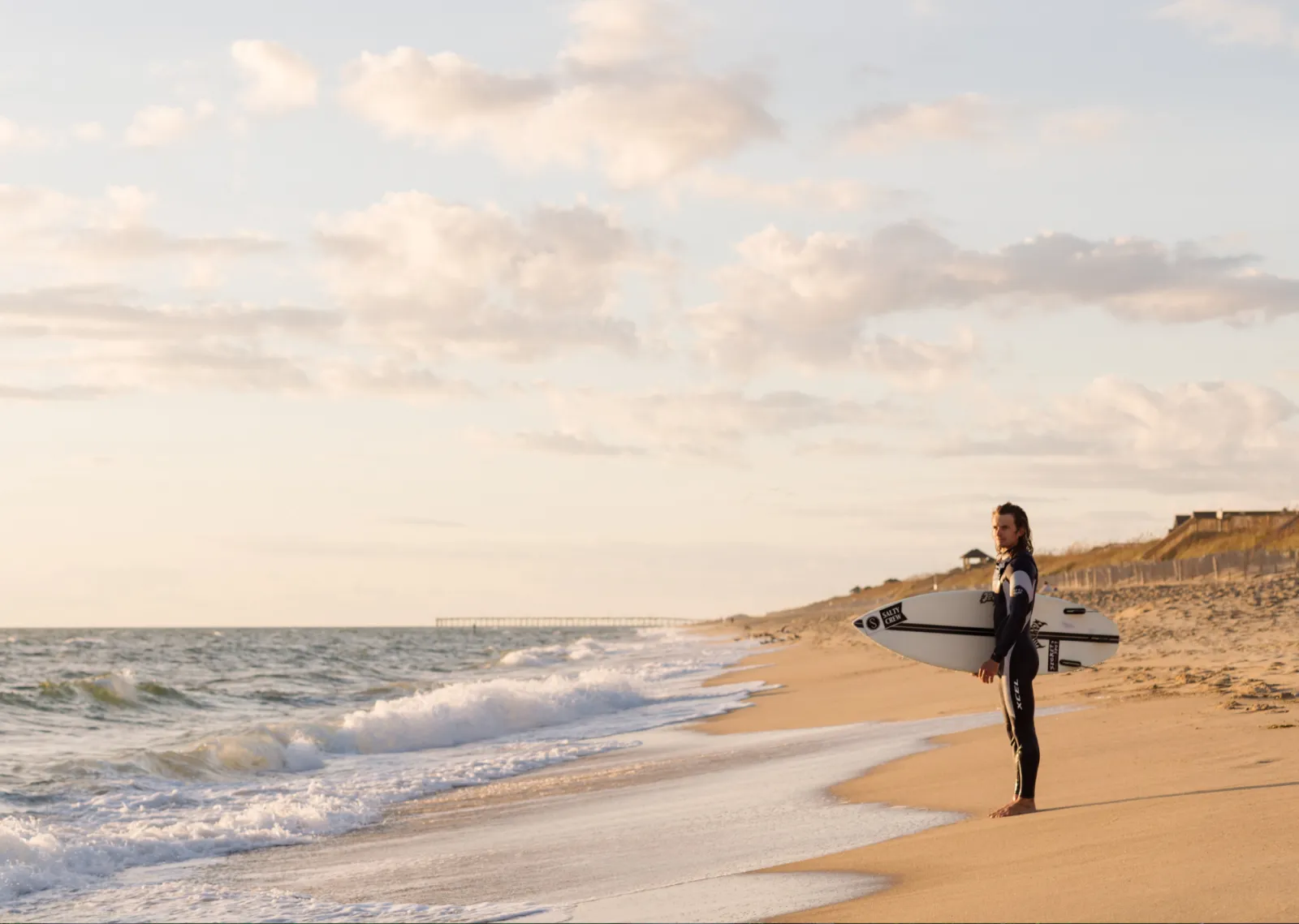 Surfer holding a board, standing on a sandy beach at sunset.