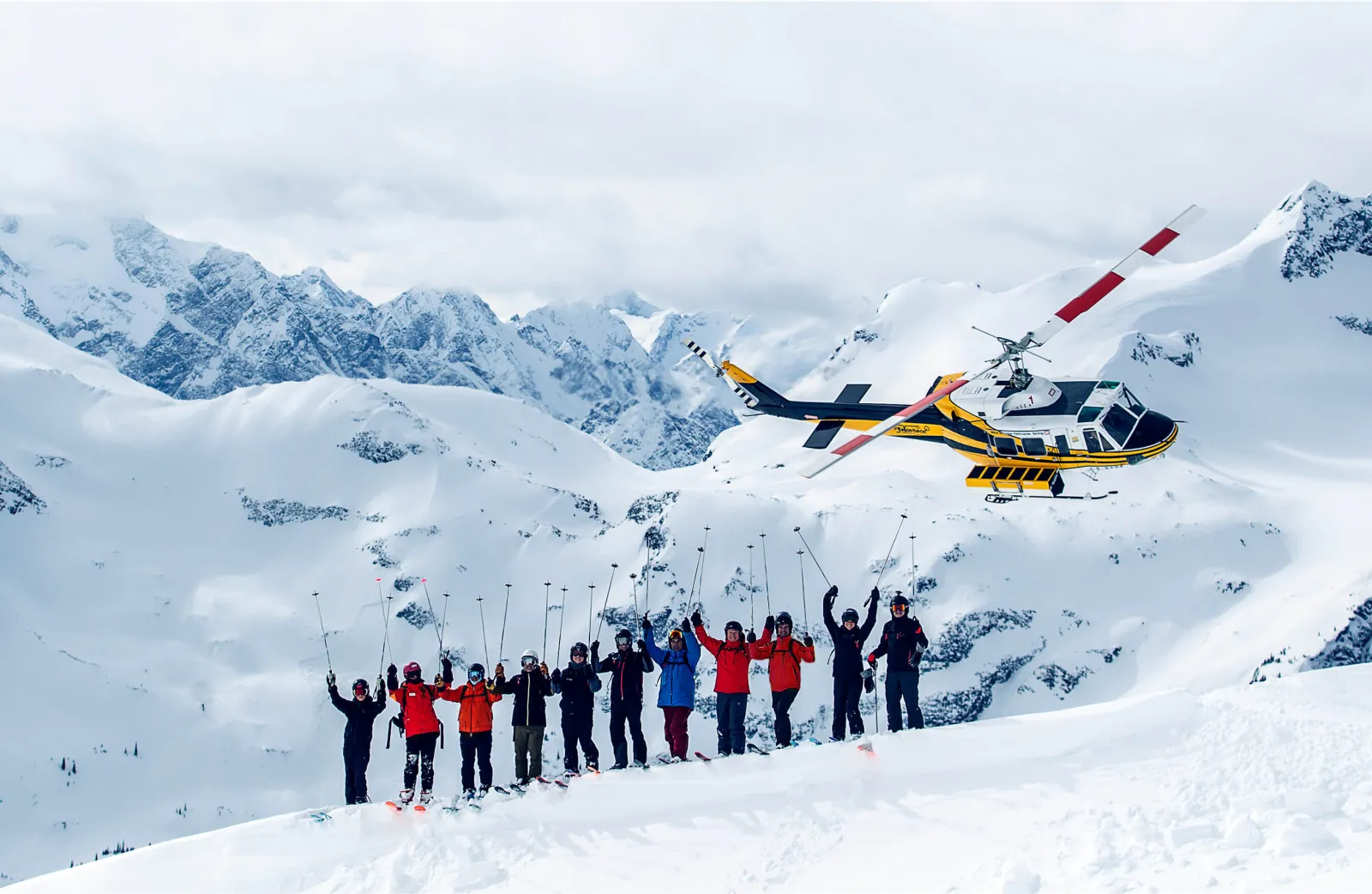 line of skiers on a ridge with their hands up, helicopter in background