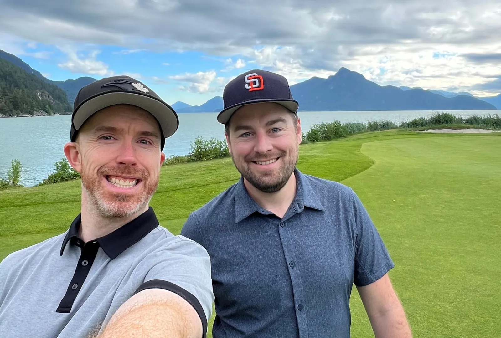 Two men smiling on a golf course by a lake, mountains in the background.