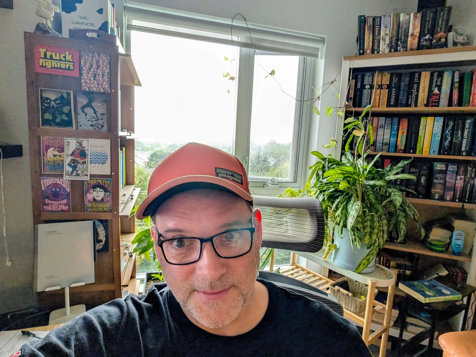 Man with glasses and cap in a home office, surrounded by books and plants.