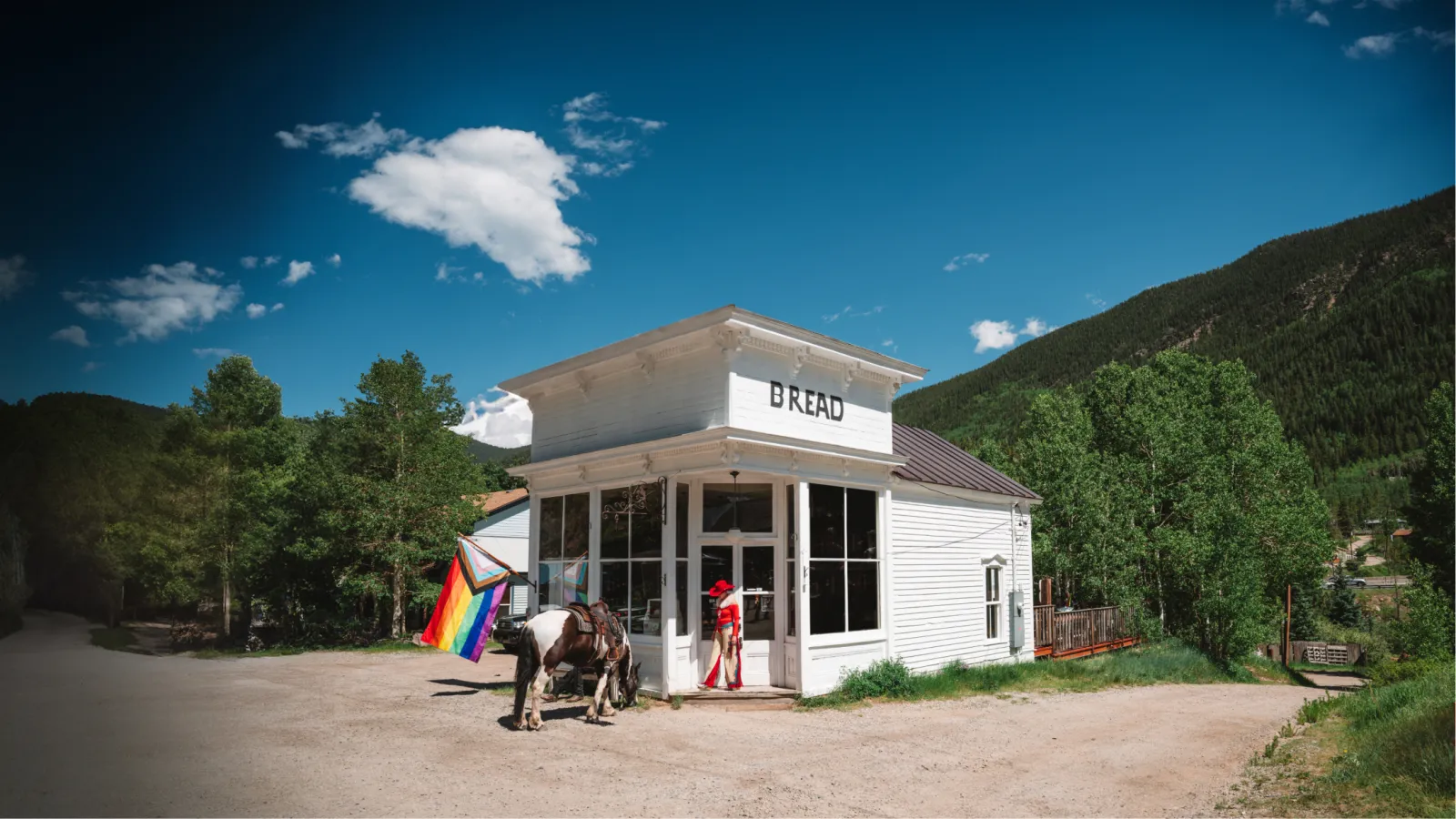 Small white building with a rainbow flag, set against a mountain backdrop and clear sky.