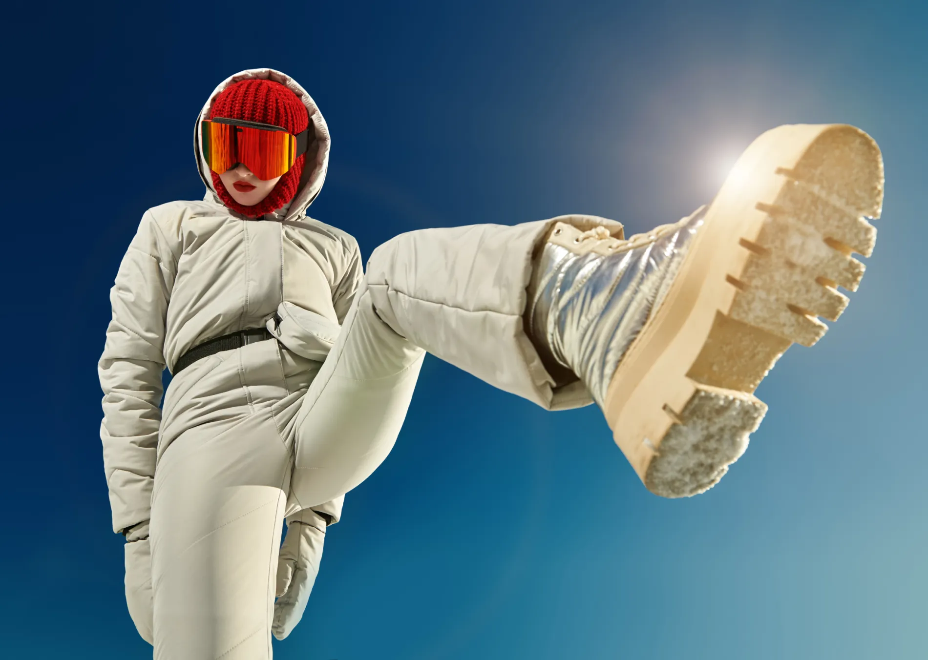 Person in winter gear kicking towards the camera under blue sky.