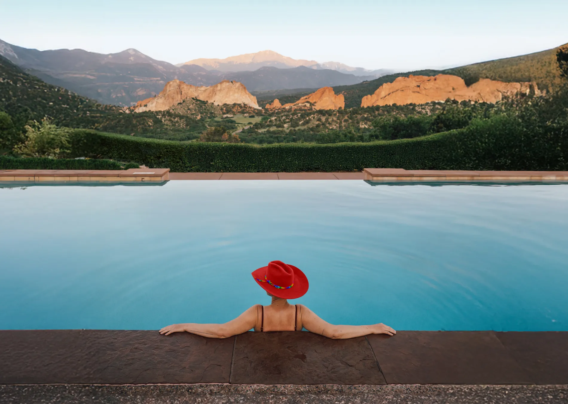 Person in a red hat relaxes in a pool, overlooking mountains at sunset.