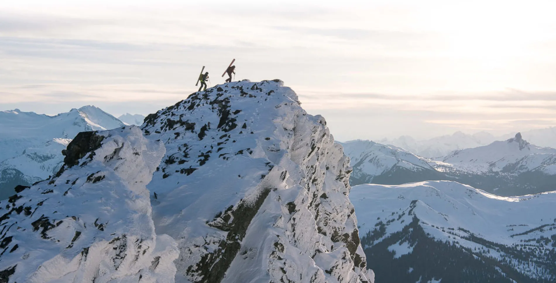 two skiers summiting a mountain at dusk with a full mountain range in the background