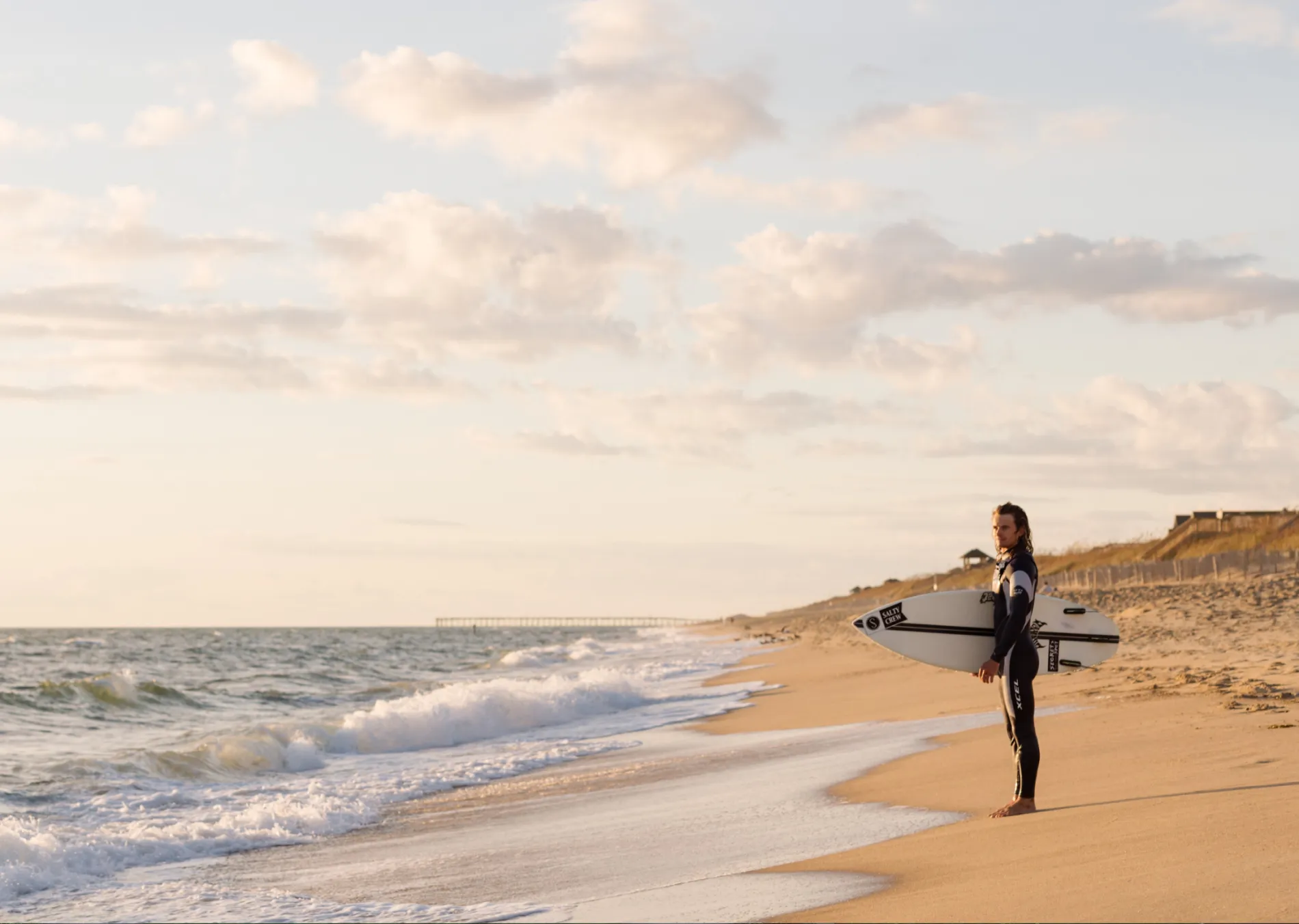 Surfer holding a board, standing on a sandy beach at sunset.